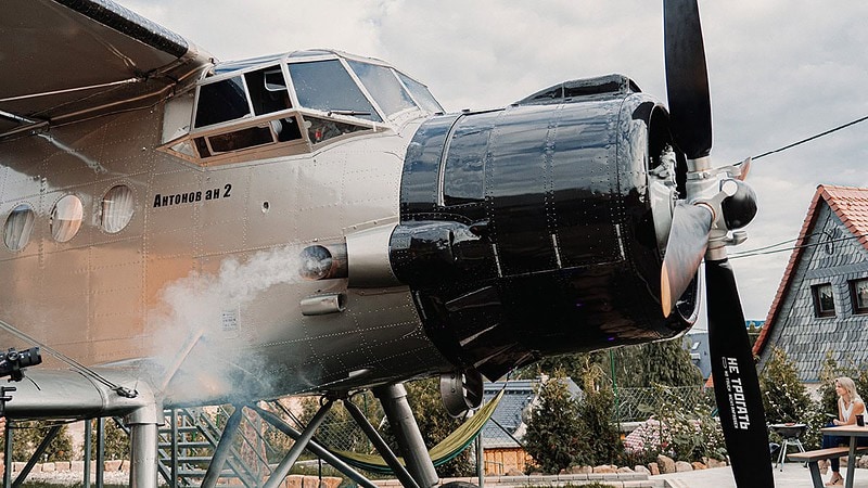 Frontansicht einer Antonov AN-2 mit großem Propeller im Gartenrestaurant, Dampf steigt auf, Besucher sitzen daneben.