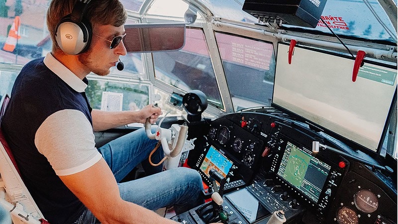 Mann mit Headset sitzt im Cockpit einer Antonov AN-2 und steuert konzentriert, viele Instrumente und Steuerhörner zu sehen.