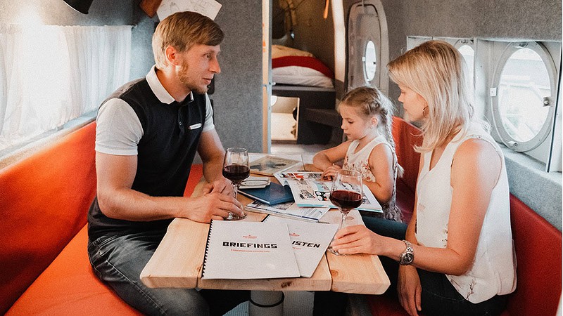Familie sitzt in der umgebauten Antonov im roten Sitzbereich an einem kleinen Tisch, Eltern trinken Wein, Kind malt.