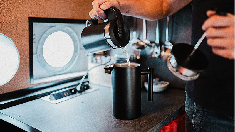 Person gießt Wasser aus einer Kanne in eine French-Press-Kaffeemaschine an einer Küchenzeile mit runden Bullaugenfenstern.