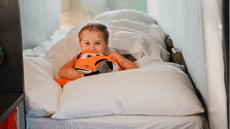 Kleines Mädchen liegt in einem schmalen Bett in der Antonov und kuschelt ein orangefarbenes Polster.