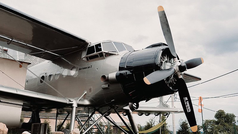 Vor dem Antonov-Flugzeug steht ein rustikales Holzfass und eine kleine Sauna mit Holztür, umgeben von Gartenwegen.