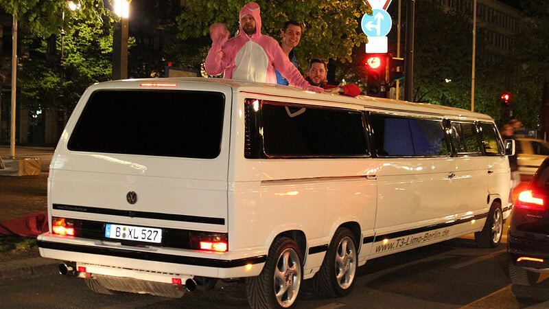 Weiße Volkswagen T3 Stretchlimousine fährt nachts, drei verkleidete Passagiere stehen lachend aus dem Dach, bunte Lichter.