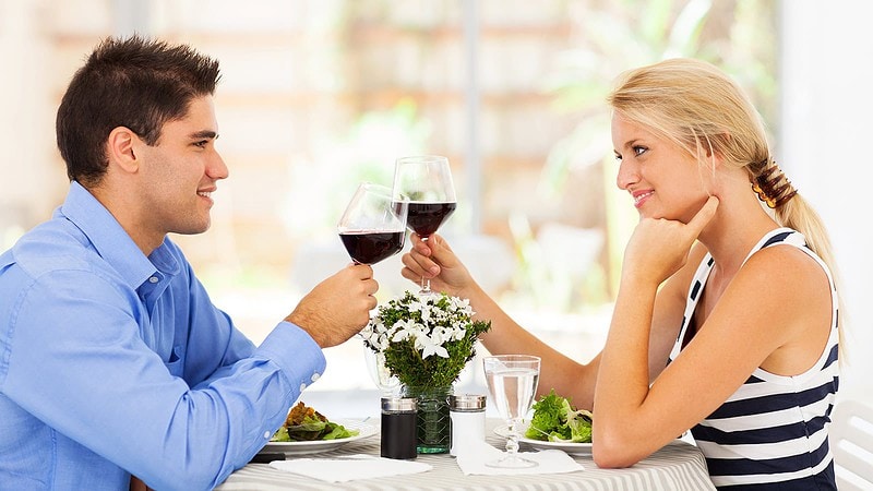Pärchen stößt beim romantischen Dinner mit Rotwein an, sitzt an einem gedeckten Tisch mit Salat und Blumen.