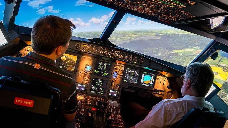 Zwei Männer steuern im A320-Cockpit-Simulator, umgeben von leuchtenden Instrumenten und einer virtuellen Landschaft.