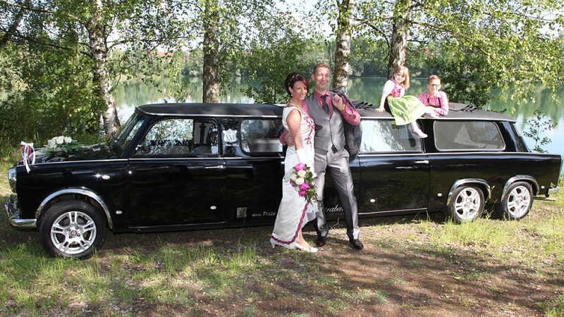 Ein Brautpaar mit Kindern lässt sich vor einer Trabant-Stretchlimousine fotografieren.