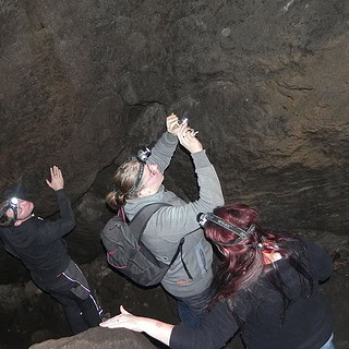 Kletterer in Helmen erklimmen mit Seil gesichert eine steile Felswand im Inneren einer dunklen Höhle.
