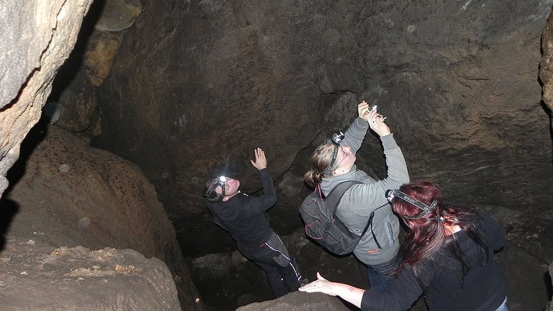 Kletterer in Helmen erklimmen mit Seil gesichert eine steile Felswand im Inneren einer dunklen Höhle.