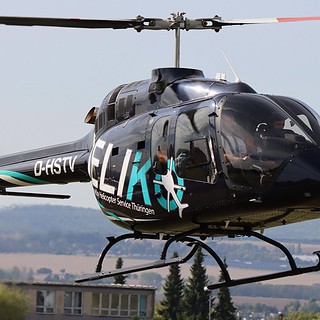 Schwarzer Helikopter mit dem Logo ELI fliegt über grüne Felder, rotierende Rotorblätter vor blauem Himmel.