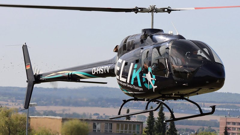Schwarzer Helikopter mit dem Logo ELI fliegt über grüne Felder, rotierende Rotorblätter vor blauem Himmel.