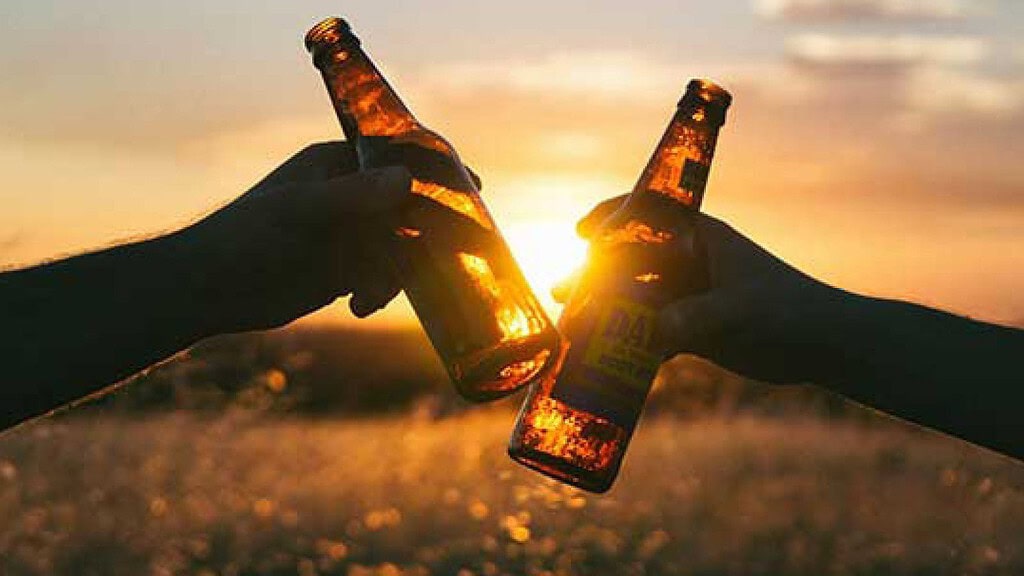 Zwei Hände stoßen im Abendlicht mit Bierflaschen an, die Sonne leuchtet durch das bernsteinfarbene Glas.