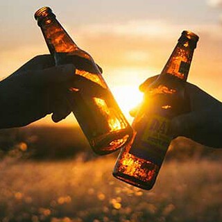 Kneipentour-Aeussere-Neustadt-in-Dresden2.jpg Zwei Hände stoßen im Abendlicht mit Bierflaschen an, die Sonne leuchtet durch das bernsteinfarbene Glas.