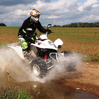 Fahrer auf weißem Quad fährt durch eine schlammige Pfütze auf einem Feldweg und spritzt Wasser auf.