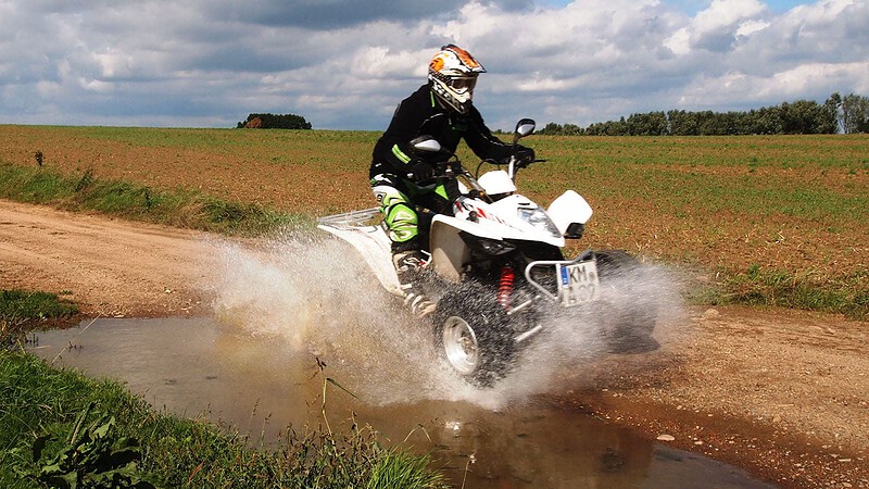 Fahrer auf weißem Quad fährt durch eine schlammige Pfütze auf einem Feldweg und spritzt Wasser auf.