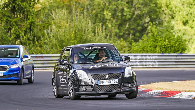 Ein schwarzer Suzuki wird von einem blauen Fahrzeug auf der Rennstrecke verfolgt.