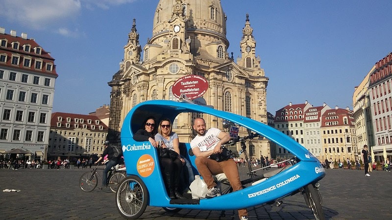 Pärchen sitzt in einer blauen Rikscha vor der barocken Frauenkirche in Dresden, Fahrer radelt durch die Stadt.