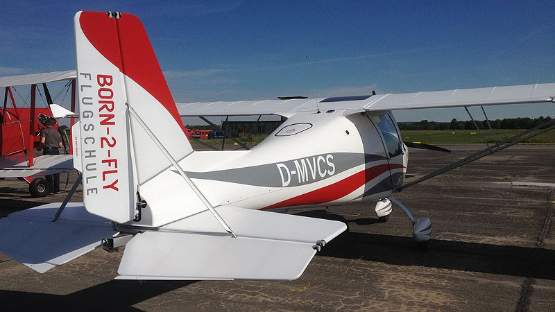 Ultraleichtflugzeug Born-2-Fly startet von der Wiese, Pilot unter Helm, Propeller dreht sich, Wälder im Hintergrund.