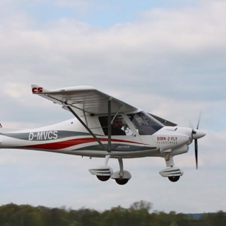 Weißes Flugzeug D-ECBX mit rotem Streifen rollt vor den Hangars, ein Mann in Shorts kontrolliert das Fahrwerk auf dem Rasen.