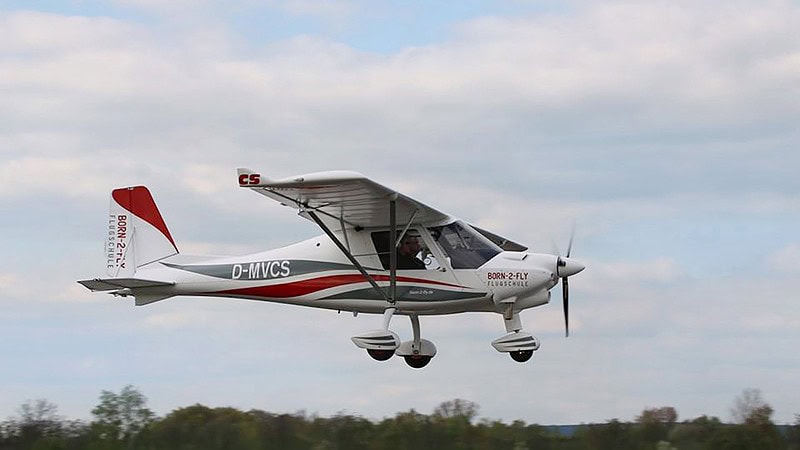 Weißes Flugzeug D-ECBX mit rotem Streifen rollt vor den Hangars, ein Mann in Shorts kontrolliert das Fahrwerk auf dem Rasen.