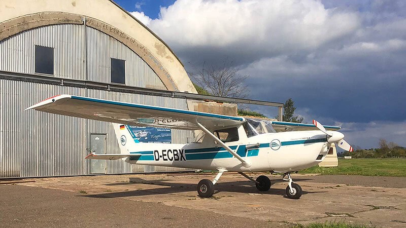 Blau-weißes Flugzeug D-EFAN steht auf einer Graspiste, nebenhangars und Bäume, Tagessonnenlicht reflektiert.