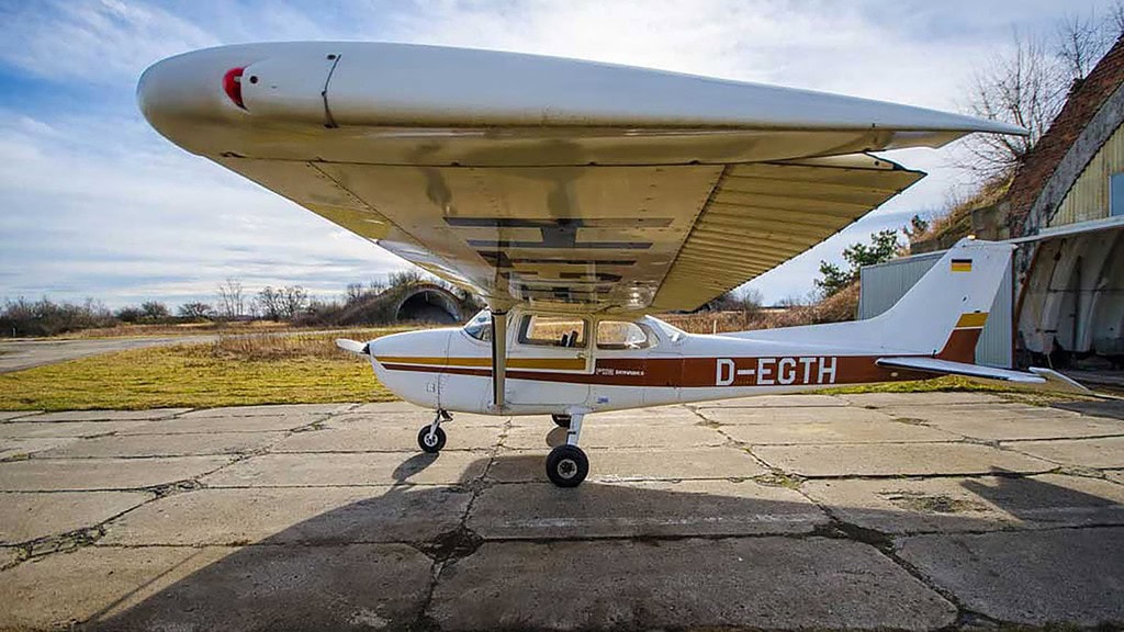 Kleine zweisitzige Maschine D-MVCS steht auf dem Flugplatz, rotes Dach und weiße Karosserie mit grünen Feldern dahinter.