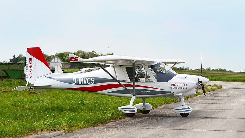 Kleines blaues Flugzeug startet in den Himmel, grüne Felder und Bäume im Hintergrund, Motorhaube glänzt.