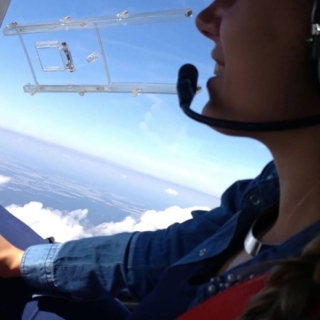 Schwarzer Hubschrauber mit gelben Streifen schwebt über weitläufige Wiesenlandschaft, Bäume und blauer Horizont.