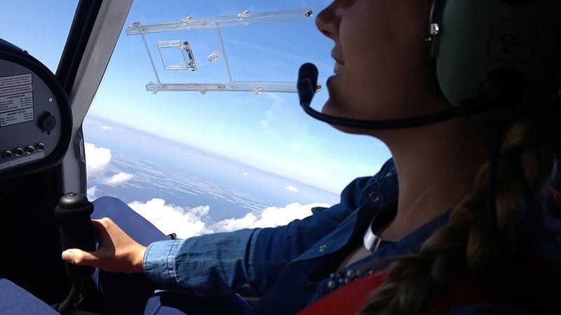 Schwarzer Hubschrauber mit gelben Streifen schwebt über weitläufige Wiesenlandschaft, Bäume und blauer Horizont.