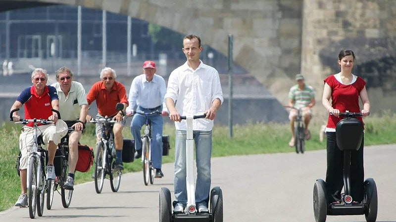 Ein Mann und eine Frau fahren in Dresden an der Elbe auf einem Segway entlang. Daneben fahren ältere Männer auf Fahrrädern.