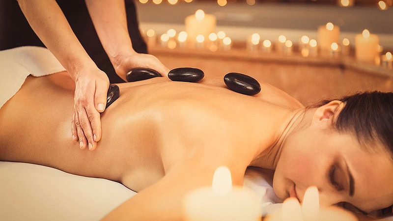 Vollkommene Entspannung bei Hot-Stone-Massage, glatte schwarze Steine liegen auf geöltem Rücken neben Kerzenlicht.