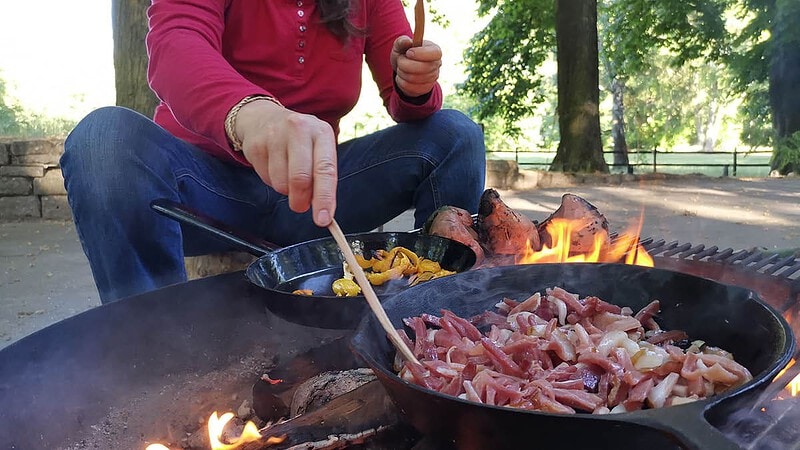 Frau am Lagerfeuer mit zwei Pfannen und Gemüse darüber