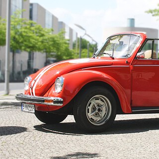 Roter VW Käfer Cabrio fährt durch eine sonnige Berliner Straße, grüne Allee und moderne Häuser im Hintergrund.