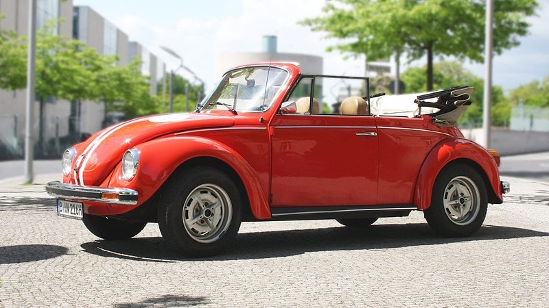 Roter VW Käfer Cabrio fährt durch eine sonnige Berliner Straße, grüne Allee und moderne Häuser im Hintergrund.