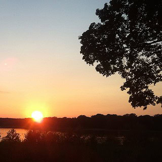 Ausblick auf einen See bei einem Sonnenuntergang. Auf dem See fährt ein Segelboot.