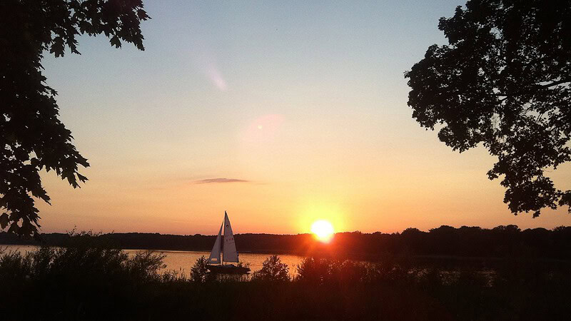 Ausblick auf einen See bei einem Sonnenuntergang. Auf dem See fährt ein Segelboot.