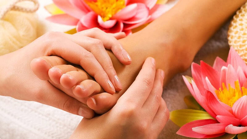 Zarte Hände massieren den Fußknöchel einer Person, nebenbei leuchtet eine rosa Blüte als Dekoration.