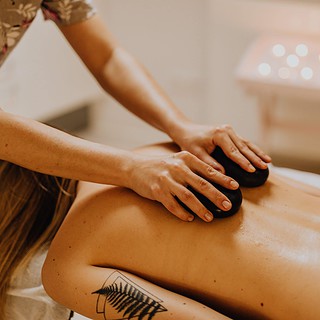 Therapeutin massiert den geölten Rücken einer liegenden Person mit zwei warmen schwarzen Massagesteinen.