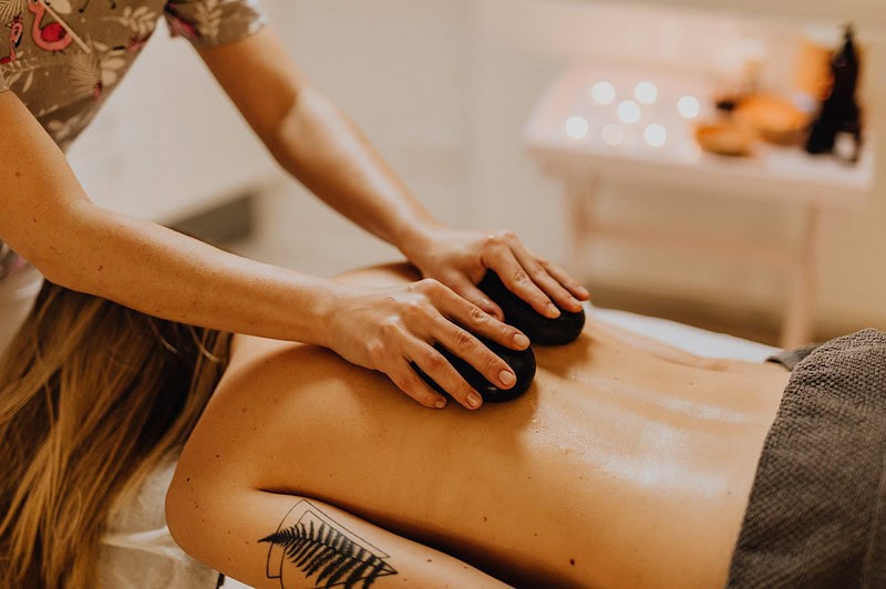 Therapeutin massiert den geölten Rücken einer liegenden Person mit zwei warmen schwarzen Massagesteinen.