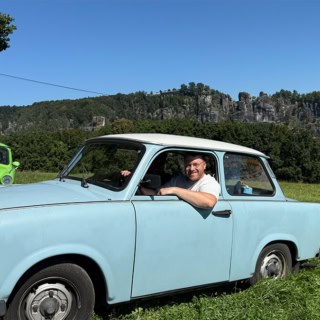 Mann lehnt sich aus dem Fenster eines blauen Trabant, hinten fährt ein grüner Trabant vorbei, Felswände im Hintergrund.