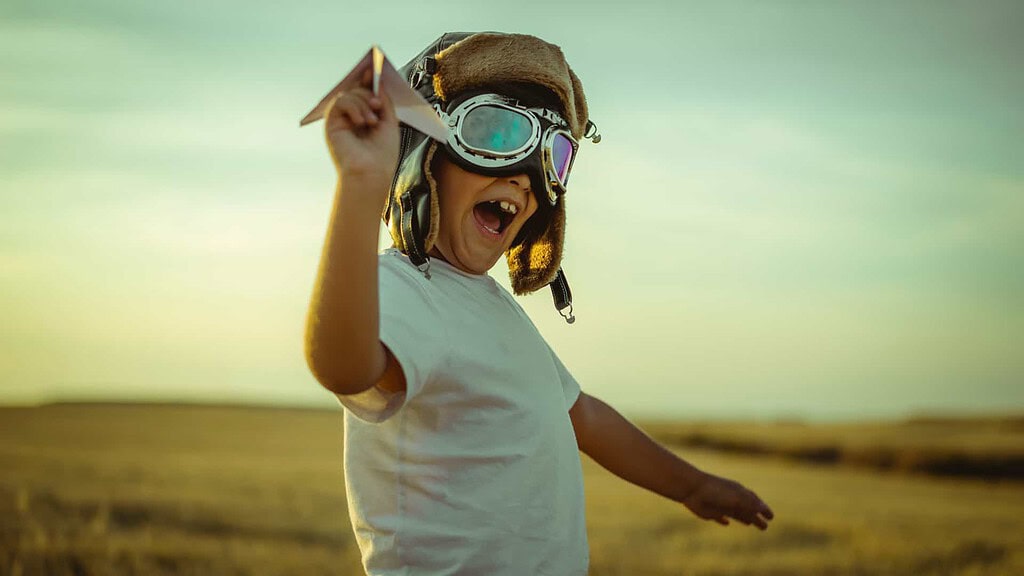 Ein glückliches Kind mit Fliegermütze und Pilotenbrille, spielt mit einem Papierflieger auf einer Wiese.