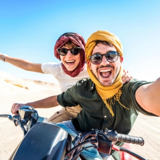 Junges Paar mit Sonnenbrillen und gelben Tüchern fährt lachend auf einem Quad durch die Wüste und macht ein Selfie.