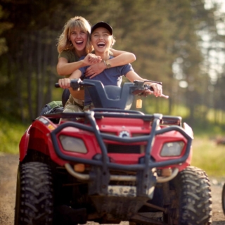 Zwei junge Frauen fahren lachend auf einem roten Quad durch einen Waldweg, Beifahrerin umarmt die Fahrerin von hinten.
