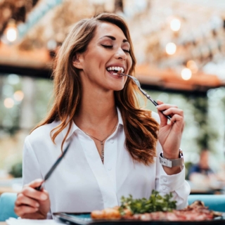 Lachende Frau in weißer Bluse genießt ihr Essen im Freien, hält eine Gabel mit Salat vor Bokeh-Lichtern.