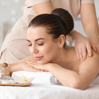 Frau liegt entspannt auf dem Bauch und erhält eine sanfte Rückenmassage, neben ihr stehen Ölfläschchen und eine weiße Blüte.