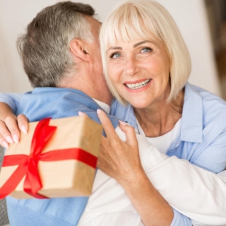Oma bekommt von Opa ein Geschenk überreicht und wird dabei umarmt.