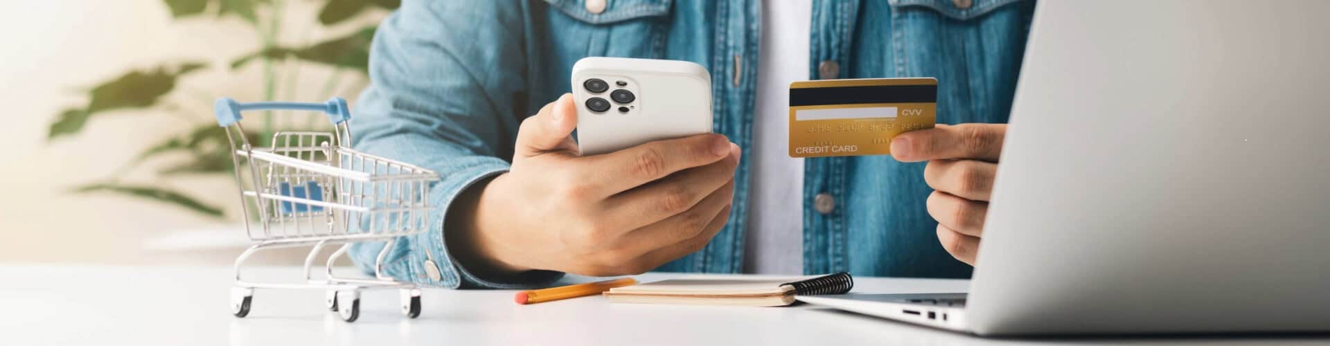 Ein Mann kauft auf einer Website ein, er hält eine Zahlungskarte in der Hand, neben ihm steht ein Miniatur-Einkaufswagen