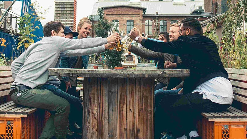 Eine Gruppe Personen stößt an einem Esstisch im freien an mit Getränken in der Hand.