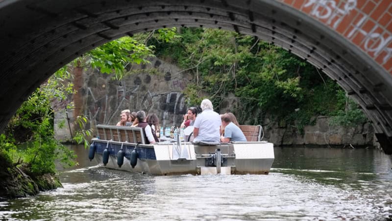Motorboot mit vier Passagieren fährt unter einer Brücke auf dem Leipziger Kanal, dichtes Grün an den Ufern.