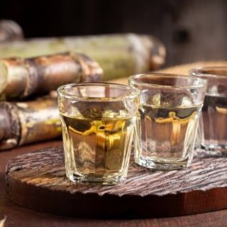 Drei mit Chachaca gefüllte Shotgläser stehen auf einem Brett aufgereiht auf einem Tisch. Im Hintergrund liegt Zuckerrohr auf dem Tisch.