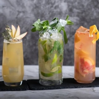 Fünf verschiedene und bunte Cocktails in unterschiedlicher Größe stehen aufgereiht auf einem Untersetzer vor einem dunkelgrauen Hintergrund.